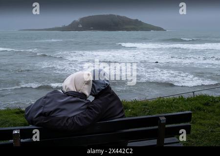 Meteo per il Regno Unito. Due persone che abbracciano e si siedono su una panchina che si affaccia sul mare verso la storica Looe Island al largo della costa della Cornovaglia nel Regno Unito. Foto Stock