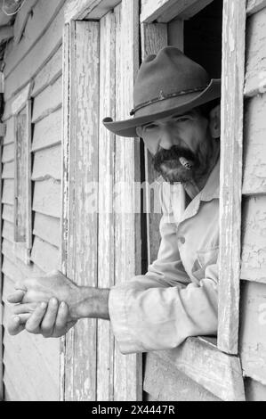 Un'immagine a infrarossi di un coltivatore di tabacco che indossa un cappello da cowboy che fuma un sigaro vicino alla finestra della sua fattoria nella Valle di Vinales, Vinales, Cuba Foto Stock