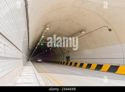 Tunnel autostradale a due corsie in cemento bianco con luci fluorescenti e frecce verdi sul soffitto e cavi elettrici che corrono lungo la parete laterale Foto Stock