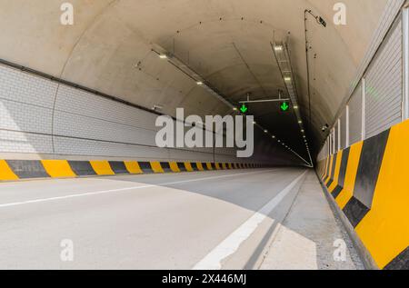 Tunnel autostradale a due corsie in cemento bianco con luci fluorescenti e frecce verdi sul soffitto e cavi elettrici che corrono lungo la parete laterale Foto Stock