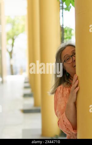 Donna più anziana con capelli bianchi e occhiali, sorridente da dietro una colonna Foto Stock