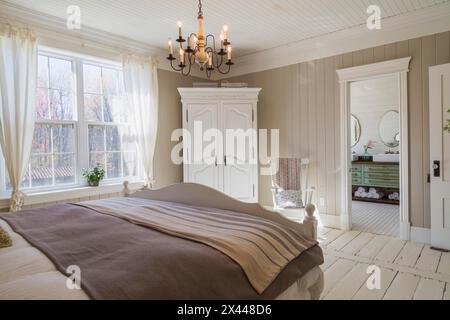 Letto king size con copriletto marrone chiaro e a strisce beige, testata e pediera in legno dipinto di bianco, sedia a dondolo, armadio alto nella padronanza Foto Stock