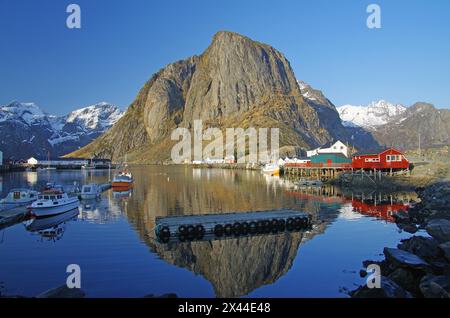 Montagne aride e case rosse che si riflettono nelle acque calme di un fiordo, inverno, Hamnoey, Lofoten, Nordland, Norvegia Foto Stock