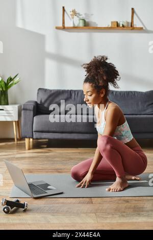 Donna riccia afro-americana in vestibilità attiva siede su un tappetino da yoga con un laptop davanti e partecipa a una sessione di fitness online. Foto Stock