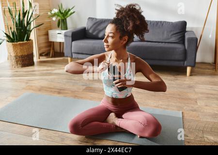 Una donna riccia afroamericana in vestibilità attiva si prende una pausa per idratarsi con una bottiglia d'acqua mentre si siede su un tappetino da yoga. Foto Stock