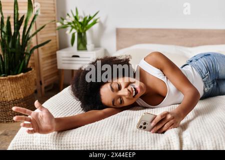 Una donna riccia afroamericana in una canotta si rilassa su un letto accanto a una vivace pianta verde in una camera da letto moderna. Foto Stock