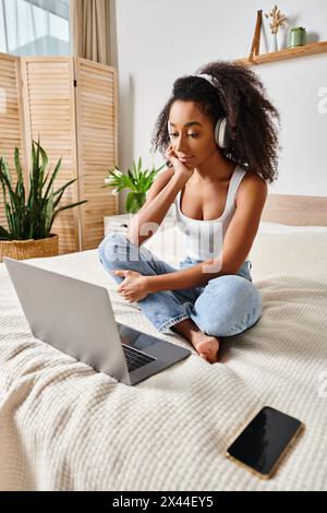 Una donna riccia afroamericana siede su un letto, concentrata sullo schermo del suo laptop in una camera da letto moderna. Foto Stock