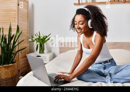 Donna riccia afroamericana in una canotta, seduta su un letto, intensamente concentrata sull'utilizzo di un computer portatile in una camera da letto moderna. Foto Stock