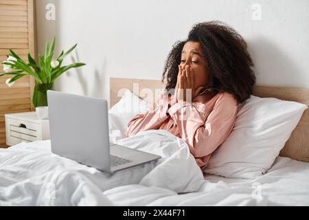 Una riccia afro-americana in pigiama giace a letto con un portatile sulle sue gambe, che si impegna con la tecnologia in un ambiente accogliente da camera. Foto Stock