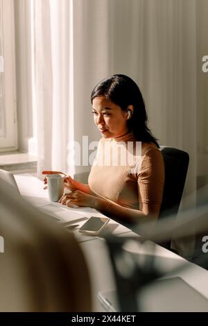 Donna d'affari sicura di sé seduta con una tazza di caffè e un computer portatile che lavora in ufficio Foto Stock