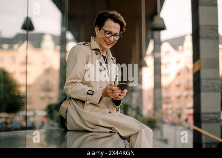 Sorridente donna anziana in pensione che usa lo smartphone mentre è seduta sulla panchina Foto Stock