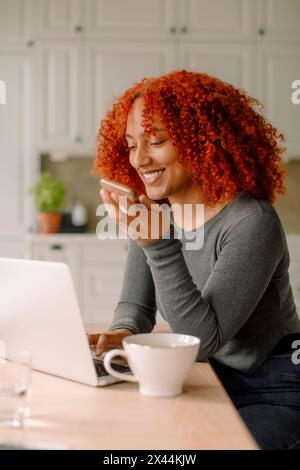 Giovane donna d'affari sorridente che parla tramite l'altoparlante dello smartphone utilizzando un computer portatile seduto sul tavolo da pranzo in cucina Foto Stock
