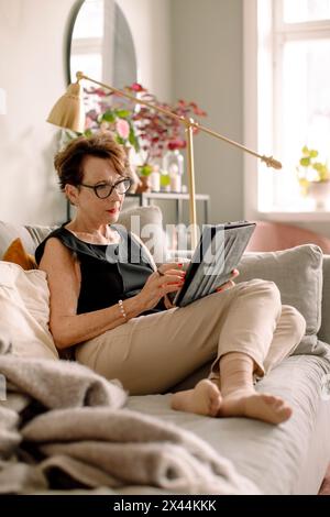 L'intera lunghezza di una donna anziana in pensione che utilizza un tablet digitale mentre si siede sul divano di casa Foto Stock