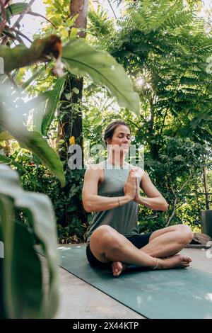 Donna matura che pratica meditazione seduto a gambe incrociate tra le piante del centro benessere Foto Stock