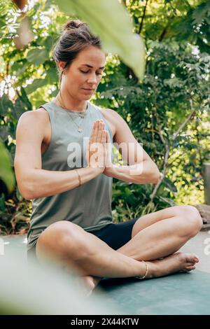 Donna matura che pratica meditazione con le mani strette mentre siede a gambe incrociate al centro benessere Foto Stock