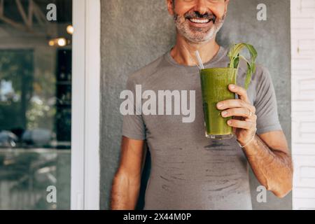 Parte centrale di un uomo sorridente che tiene un bicchiere di fresco frullato verde contro le pareti del resort Foto Stock