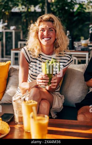 Ritratto di una donna bionda sorridente che regge un bicchiere di frullato mentre si siede al resort Foto Stock