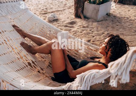 Donna che legge un libro sdraiato sull'amaca in vacanza in spiaggia Foto Stock