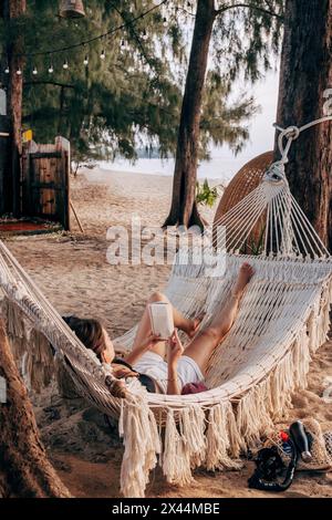 Donna che legge un libro mentre sdraiata sull'amaca in vacanza in spiaggia Foto Stock