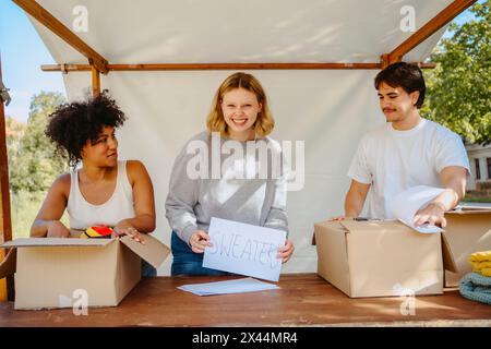 Ritratto di volontario felice con un cartello con i colleghi di sesso maschile e femminile vicino al tavolo durante un viaggio di beneficenza al communit Foto Stock