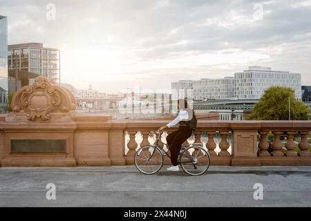 Vista laterale di una giovane donna d'affari professionista in bicicletta sul marciapiede in città contro il cielo Foto Stock