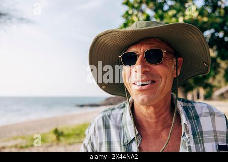 Un anziano sorridente che indossa occhiali da sole e cappello in spiaggia in vacanza Foto Stock