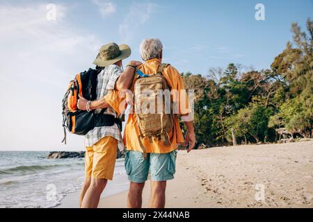 Vista posteriore ad angolo basso della coppia gay senior con zaini in piedi in spiaggia in vacanza Foto Stock