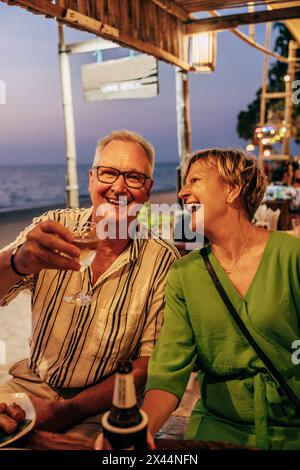 Ritratto di un anziano sorridente che si gusta il vino con una donna seduta al ristorante sulla spiaggia Foto Stock