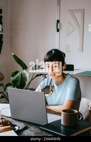 Giovane freelance che parla con un telefono con vivavoce mentre è seduta alla scrivania con un computer portatile a casa Foto Stock