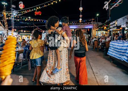 Vista posteriore della donna che porta il figlio mentre cammina con le figlie al mercato alimentare Foto Stock