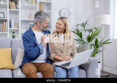 Una coppia allegra e matura si diverte a trascorrere un po' di relax insieme utilizzando un computer portatile nel loro soggiorno elegantemente arredato, trasudando felicità e comfort. Foto Stock