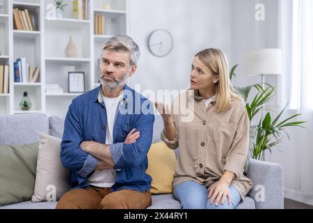 Un uomo e una donna maturi che hanno una conversazione seria in un salotto luminoso e moderno. L'uomo è sconvolto, piegando le braccia, mentre la donna sembra spiegare qualcosa. Foto Stock