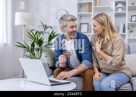 Un'affettuosa coppia matura si diverte a fare shopping online nel suo accogliente salotto, illustrando amore, tecnologia e consumismo. Foto Stock