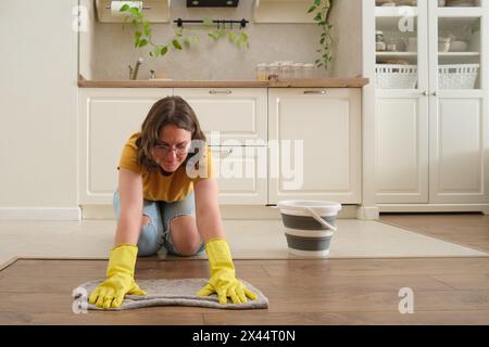 Una donna lava il pavimento con le mani in una cucina domestica, lavoro manuale. Le mani delle donne indossano guanti gialli mentre puliscono la cucina Foto Stock