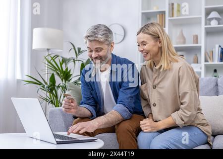 Una coppia matura e felice ama fare shopping online insieme nel suo accogliente soggiorno, dimostrando convenienza e uso tecnologico nella vita domestica moderna. Foto Stock