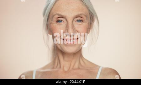 Ritratto di una donna anziana che guarda la fotocamera, sorridente. Signora anziana con capelli grigi naturali, occhi blu. Il concetto di eleganza vecchia età per la cura della pelle, la bellezza, il prodotto cosmetico con sfondo beige isolato Foto Stock