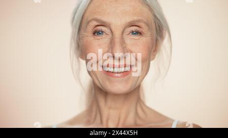 Ritratto di una donna anziana che guarda la fotocamera, sorridente. Signora anziana con capelli grigi naturali, occhi blu. Il concetto di eleganza vecchia età per la cura della pelle, la bellezza, il prodotto cosmetico con sfondo beige isolato Foto Stock