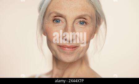 Ritratto di una donna anziana che guarda la fotocamera, sorridente. Signora anziana con capelli grigi naturali, occhi blu. Il concetto di eleganza vecchia età per la cura della pelle, la bellezza, il prodotto cosmetico con sfondo beige isolato Foto Stock