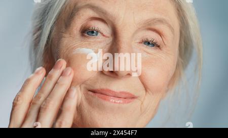 Primo piano Ritratto di una bella donna anziana che applica delicatamente la crema viso sotto gli occhi. La signora anziana rende la sua pelle morbida, liscia e senza rughe con cosmetici anti-età naturali. Prodotto per la bellezza della pelle Foto Stock