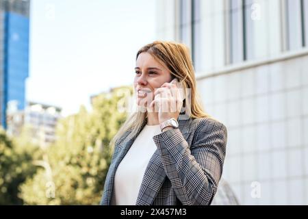 Donna d'affari in tuta che parla al telefono mentre lascia l'ufficio Foto Stock