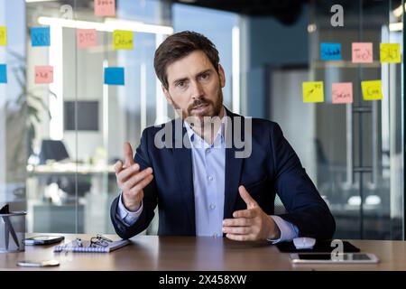 Uomo d'affari concentrato in tuta che comunica tramite videochiamata in un ufficio moderno, discutendo argomenti importanti mentre gesti con la mano. Foto Stock