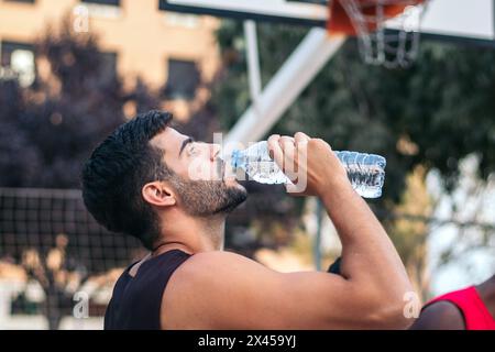 Giovane che beve acqua sul campo da pallacanestro dopo aver fatto sport e giocato a una partita Foto Stock