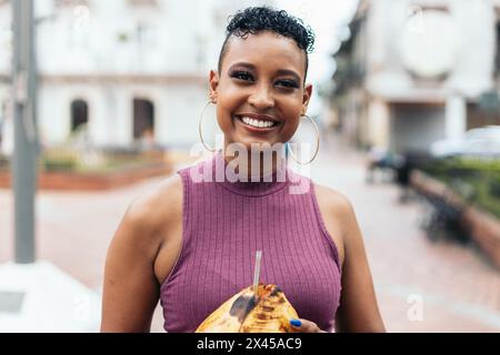 Ritratto di una donna latinoamericana corta dai capelli ricci con grandi orecchini che reggono una noce di cocco. Guarda la telecamera sorridendo e trasmette gioia e cal Foto Stock