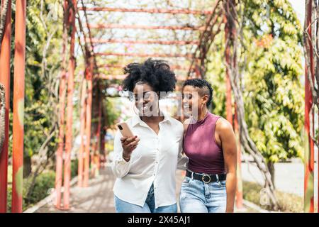 Donne nere dell'america Latina amici alla moda che si divertono a camminare in un parco e a usare lo smartphone Foto Stock