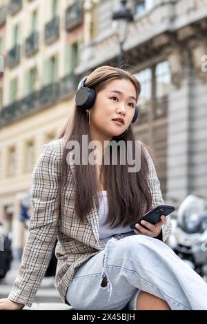 Giovane donna asiatica seduta su una panchina in una città europea che aspetta qualcuno mentre guarda lo smartphone e ascolta musica con le cuffie Foto Stock