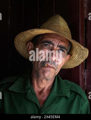 Un coltivatore di tabacco che indossa un cappello di paglia che fuma un sigaro davanti alla porta d'ingresso della sua fattoria nella Val di Vinales, Vinales, Cuba. Foto Stock
