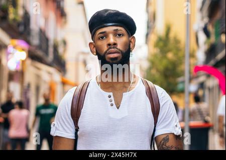 Ritratto di un elegante uomo nero con berretto e tatuaggi in piedi per la strada della città guardando la fotocamera Foto Stock