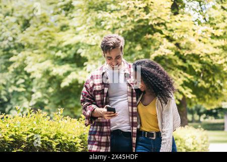 Felice giovane coppia multirazziale di uomo caucasico e donna afroamericana che fa una passeggiata nel parco mentre usa lo smartphone e ride insieme Foto Stock