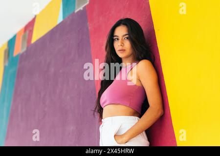 Primo piano di una giovane latina in abiti estivi appoggiata su un muro colorato e che guarda la fotocamera Foto Stock