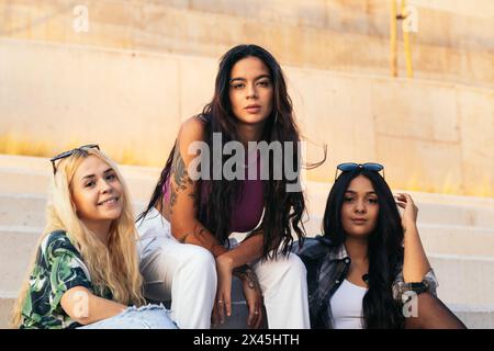 Ritratto di tre giovani donne alla moda con occhiali da sole seduti su alcuni gradini all'aperto in città. Gen-z amiche di sesso femminile che frequentano il pomeriggio Foto Stock